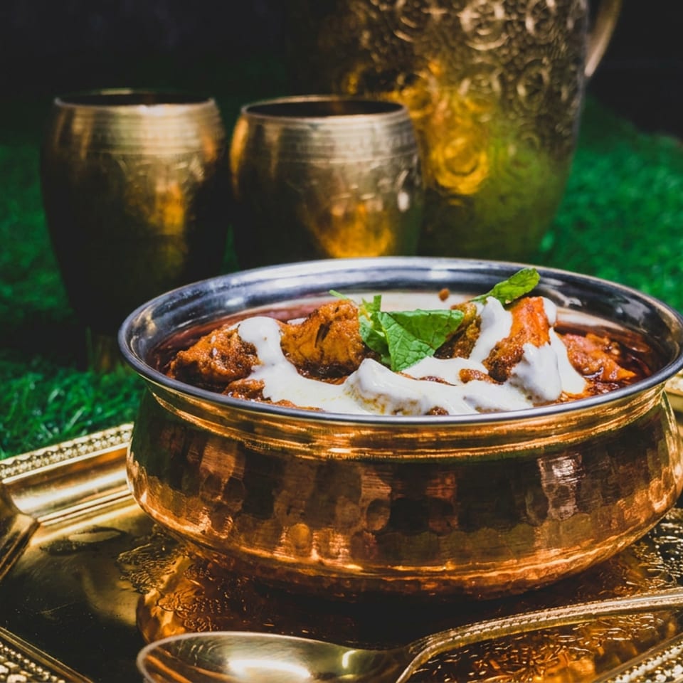 Spiced orange curry topped with yogurt and mint in a copper bowl on an ornate tray, with brass cups behind.