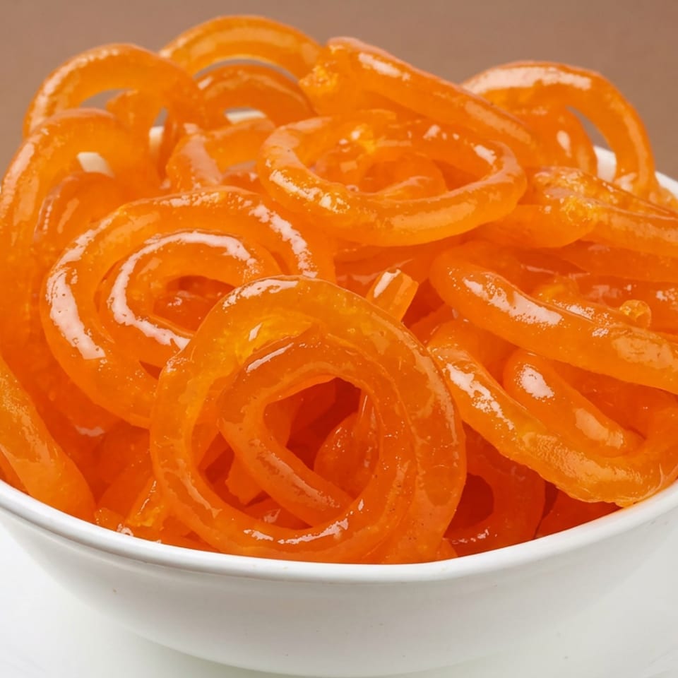 White bowl filled with glossy orange jalebi spirals on a matching plate against a brown background.
