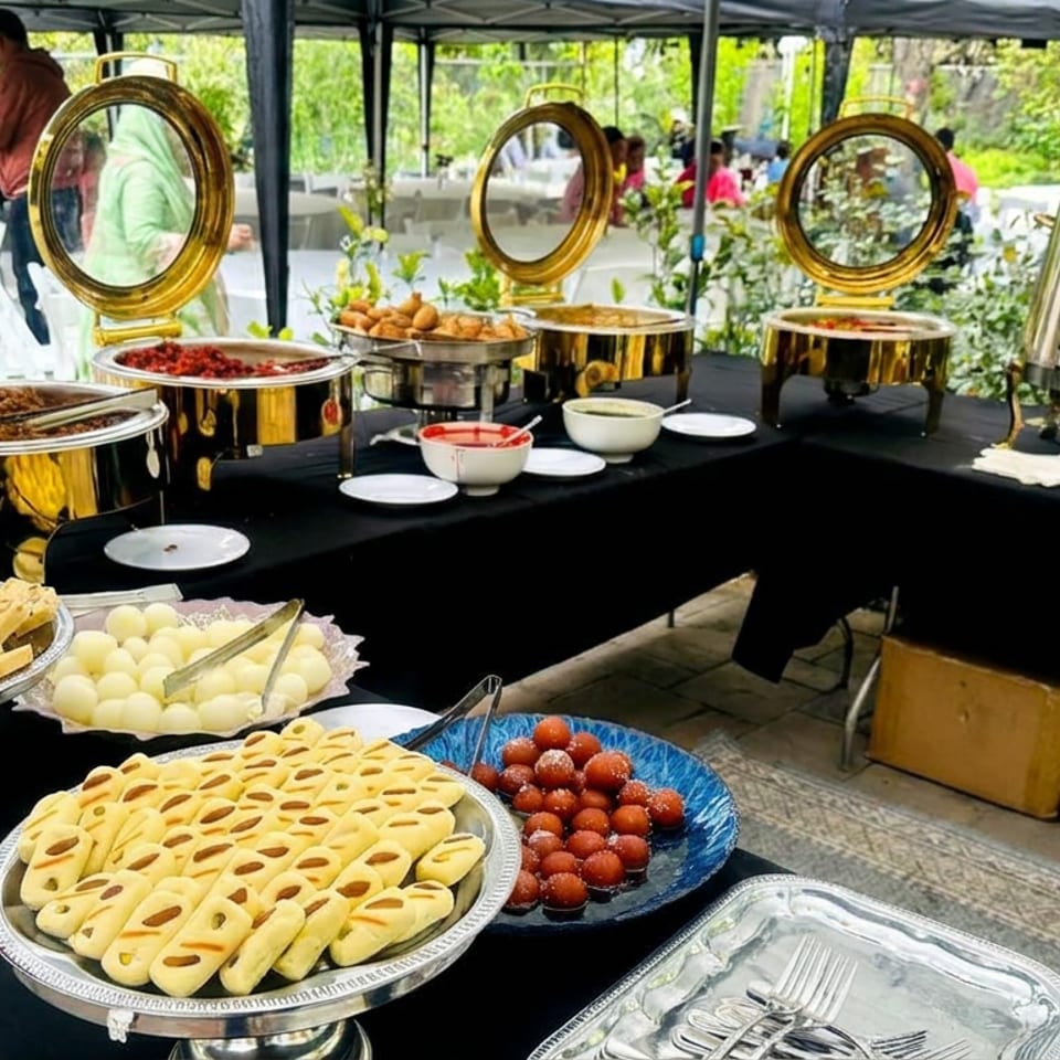 Outdoor buffet table displaying golden chafing dishes, pastries, cheese, strawberries, serving utensils, and floral decorations.