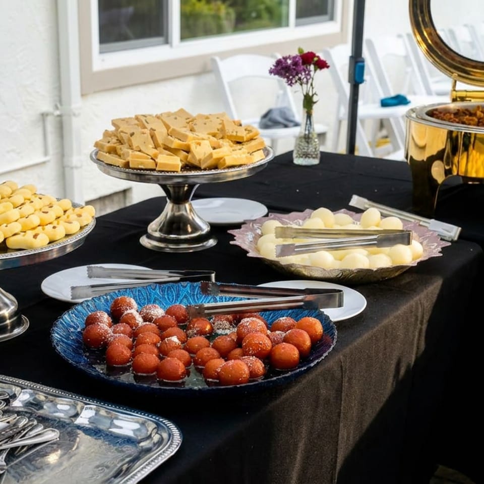 Outdoor dessert buffet with pastries, cheese squares, round sweets, serving utensils, and a gold chafing dish on a black tablecloth.